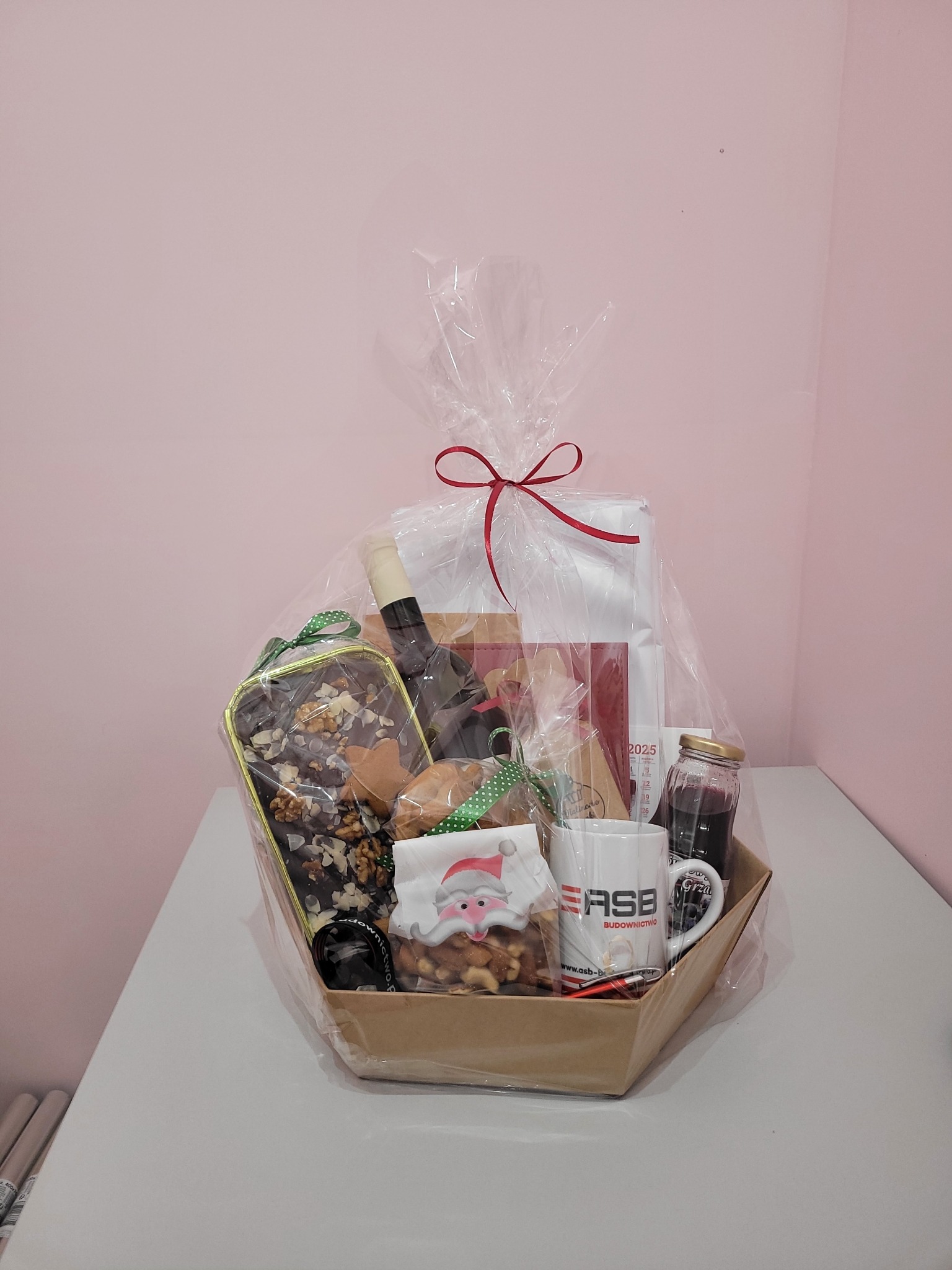 Świąteczny kosz prezentowy ze słodkościami, winem i kawą, zapakowany w celofan z czerwoną kokardą. Idealny upominek ze Świdnicy na Boże Narodzenie.