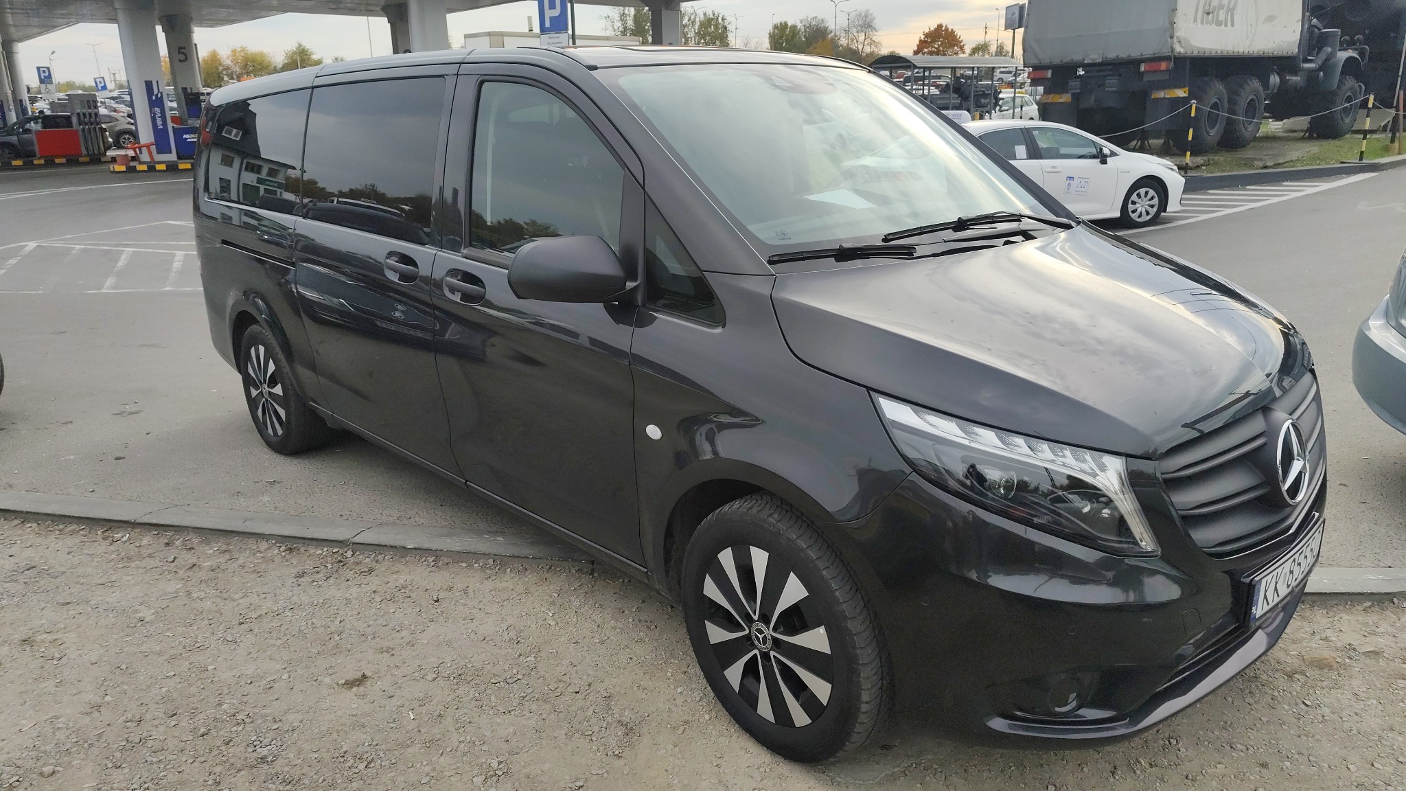 Czarny bus Mercedes Vito na parkingu stacji benzynowej, widok z boku, z widocznym fragmentem tablicy rejestracyjnej. W tle inne pojazdy i dystrybutory paliw.
