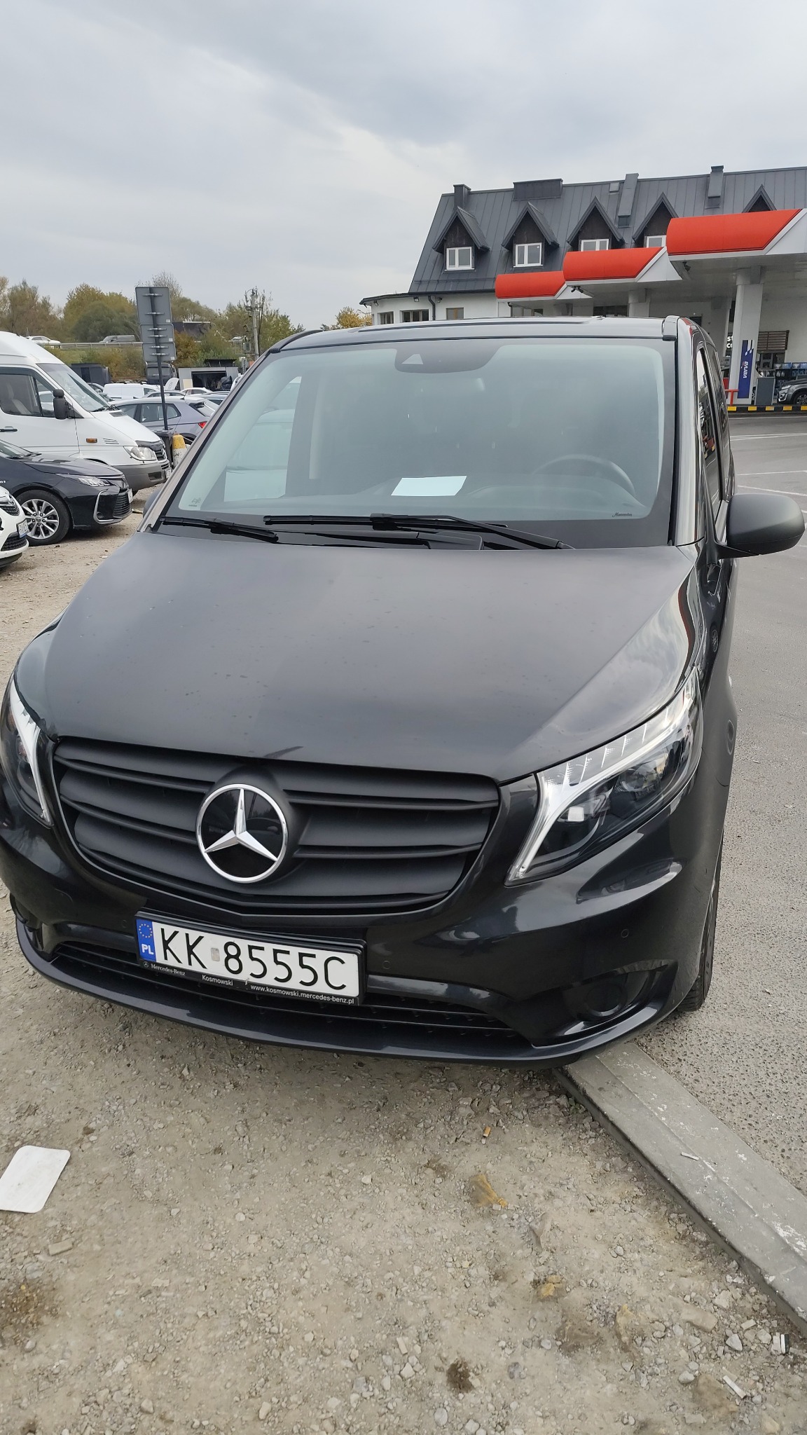 Czarny Mercedes Vito z polską rejestracją KK 8555C na parkingu, widok z przodu, w tle budynek stacji benzynowej i inne pojazdy. Pochmurny dzień.