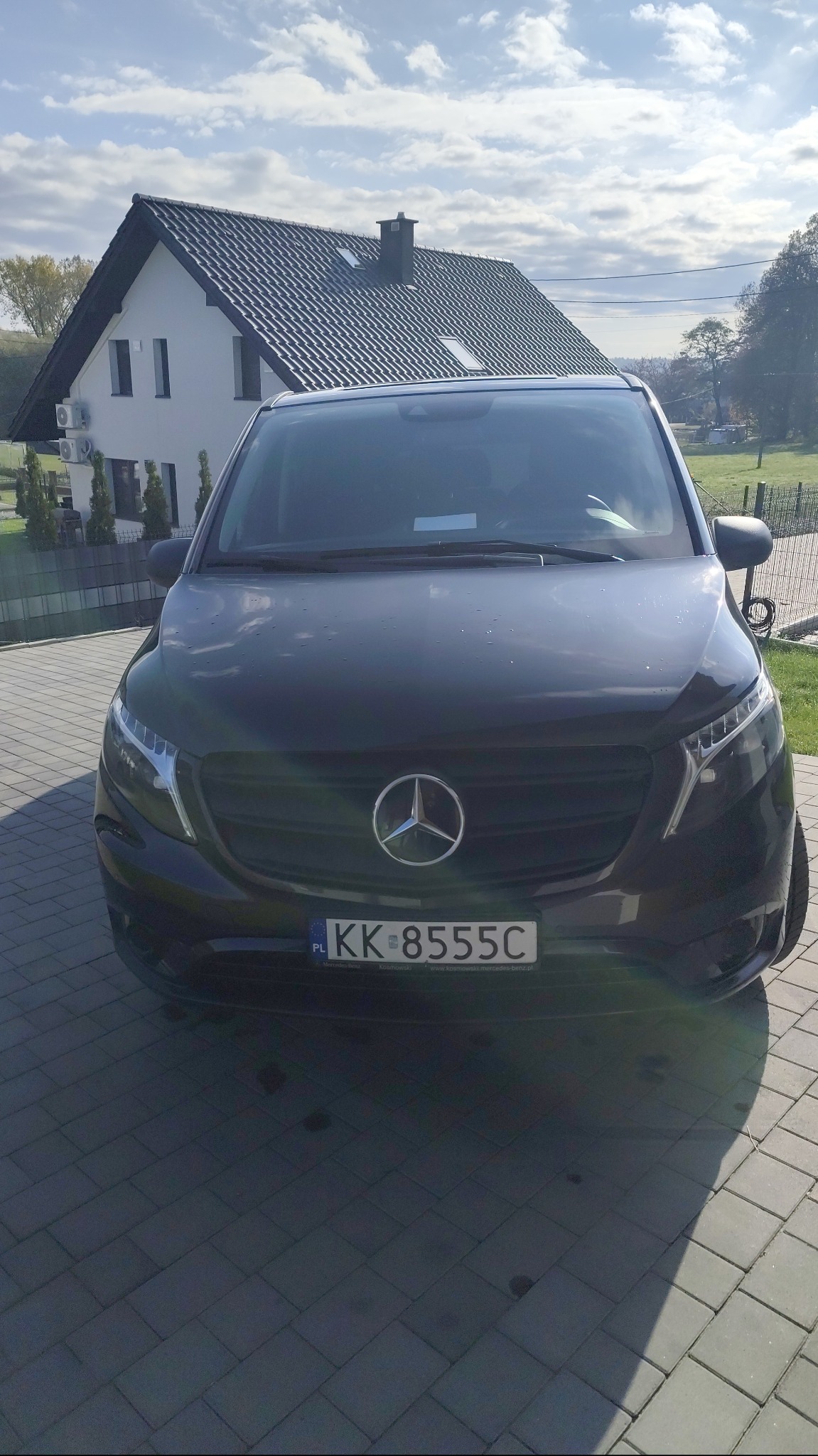 Czarny Mercedes V-klasa z numerem rejestracyjnym KK-8555C na tle domu z ciemnym dachem i błękitnego nieba. Widok z przodu, lekko z dołu.