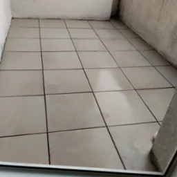 Balkon z beżowymi płytkami po umyciu. Widoczne ślady po wodzie na czystej podłodze. Ściany w jasnym odcieniu. Ujęcie z dołu, z widocznym fragmentem ramy okiennej.