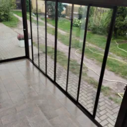 Szklana zabudowa tarasu z widokiem na ogród, po umyciu. Czyste szyby odbijają otoczenie, a na podłodze z płytek widoczne są delikatne refleksy światła.