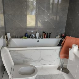 Anna Perfect - Nowoczesna łazienka z szarymi płytkami imitującymi marmur, białą wanną z kosmetykami i toaletą z otwartą klapą. Dywanik w kolorze szarym.