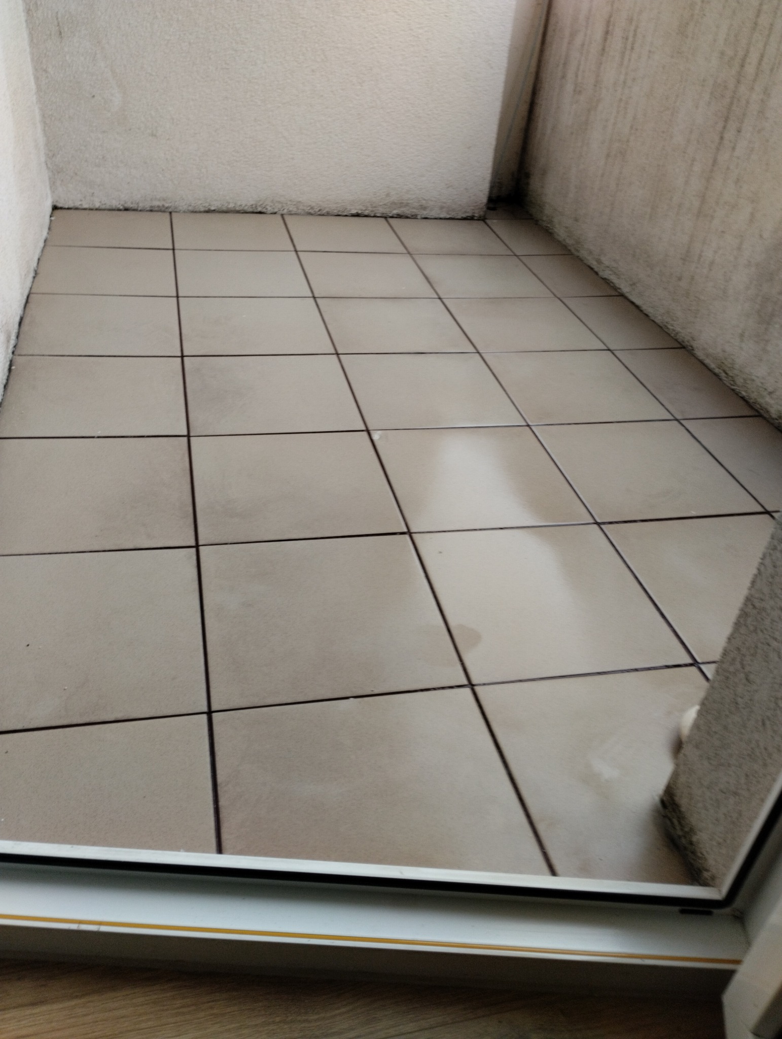 Balkon z beżowymi płytkami po umyciu. Widoczne ślady po wodzie na czystej podłodze. Ściany w jasnym odcieniu. Ujęcie z dołu, z widocznym fragmentem ramy okiennej.