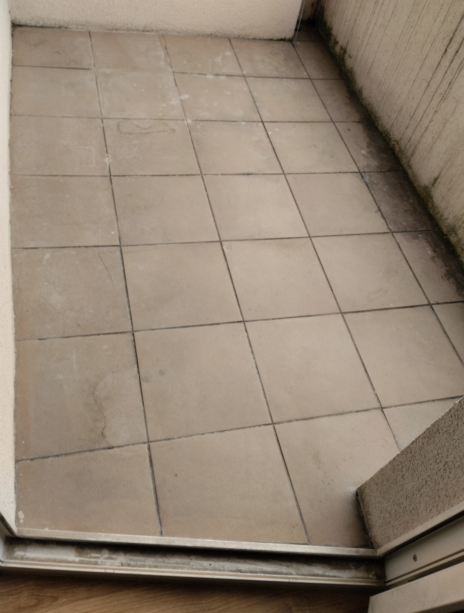Zaniedbany balkon z brudnymi, beżowymi płytkami i śladami wilgoci na ścianach. Widoczne zabrudzenia w fugach i przy krawędziach. Perspektywa z góry.