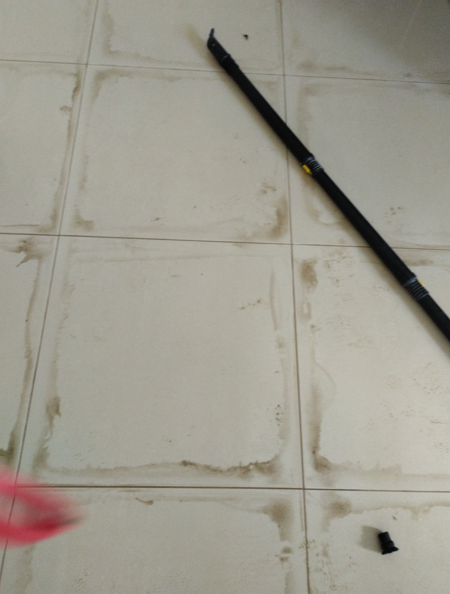 Białe, brudne płytki podłogowe z czarną rurą od odkurzacza i odczepioną końcówką. Widoczne ślady po zalaniach i zabrudzeniach w fugach.