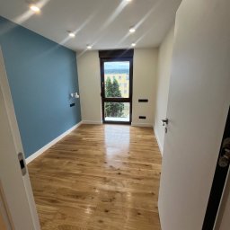 Yevhen Zatula - Wnętrze pokoju po remoncie: świeżo pomalowana niebieska ściana, jasna drewniana podłoga, białe ściany i drzwi, okno z widokiem na zieleń. Nowoczesne oświetlenie.