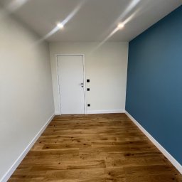 Yevhen Zatula - Wnętrze po remoncie: drewniana podłoga, białe ściany z drzwiami i fragment niebieskiej ściany. Widoczne oświetlenie sufitowe.