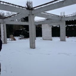 Yevhen Zatula - Surowa konstrukcja domu w zimowej scenerii: betonowe słupy i belki zbrojeniowe na tle śnieżnego krajobrazu. Częściowo wymurowane ściany z białych bloczków.