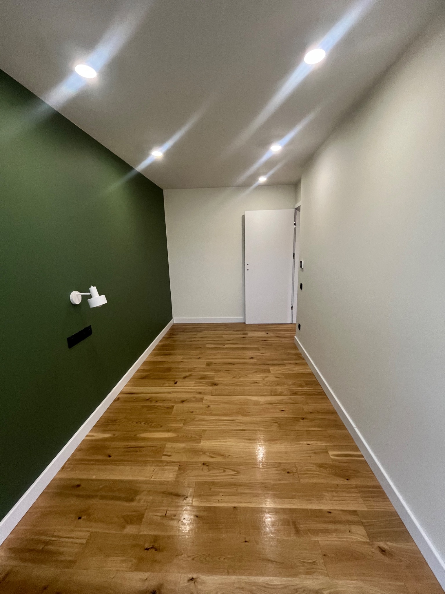 Korytarz po remoncie: drewniana podłoga, ściana w kolorze butelkowej zieleni z lampką, biała ściana z drzwiami. Widoczne oświetlenie sufitowe.