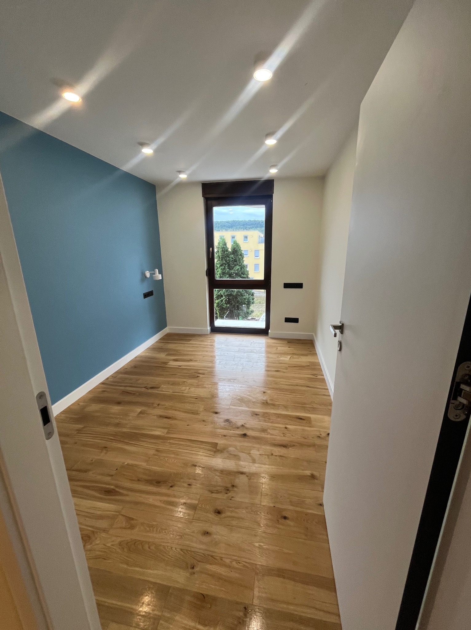 Wnętrze pokoju po remoncie: świeżo pomalowana niebieska ściana, jasna drewniana podłoga, białe ściany i drzwi, okno z widokiem na zieleń. Nowoczesne oświetlenie.