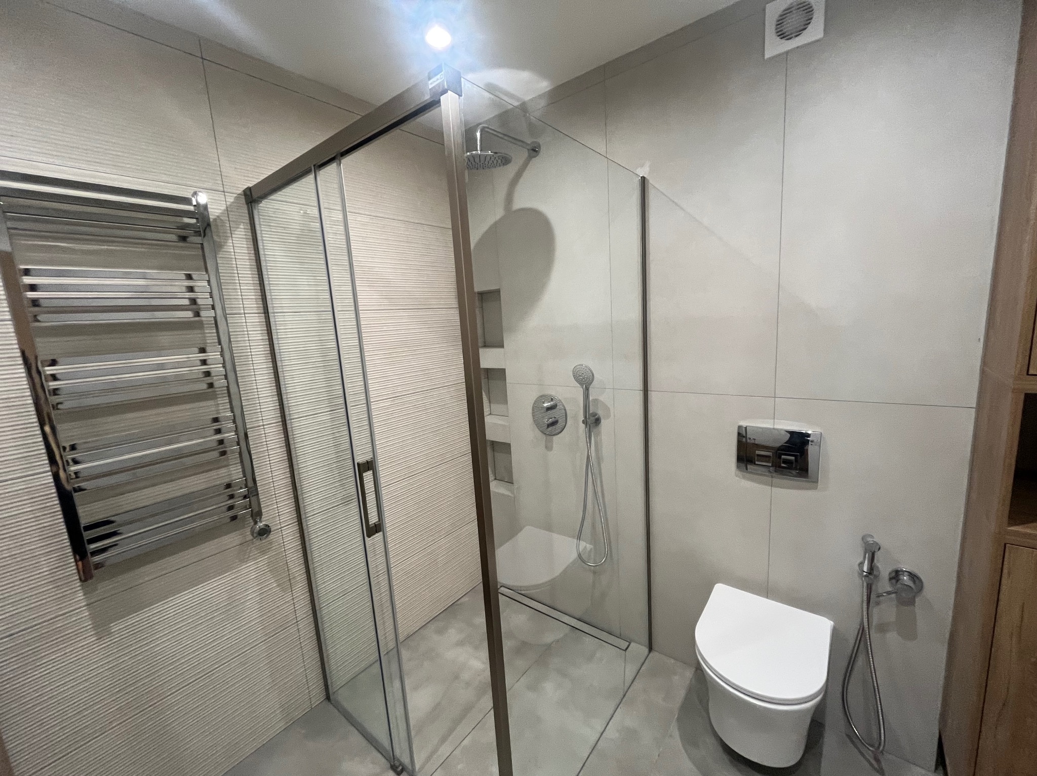 Nowoczesna łazienka z kabiną prysznicową typu walk-in, grzejnikiem drabinkowym i podwieszaną toaletą. Minimalistyczny design, szare płytki z subtelną fakturą.