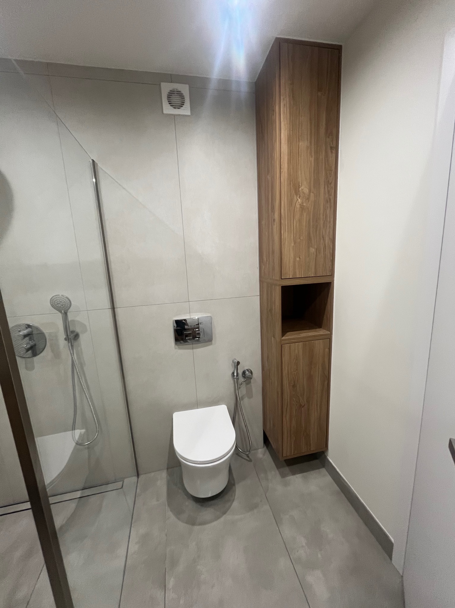 Nowoczesna łazienka z kabiną prysznicową, podwieszaną toaletą i wysoką szafką z drewna. Szare płytki podłogowe i ścienne, minimalistyczny design, oświetlenie sufitowe.