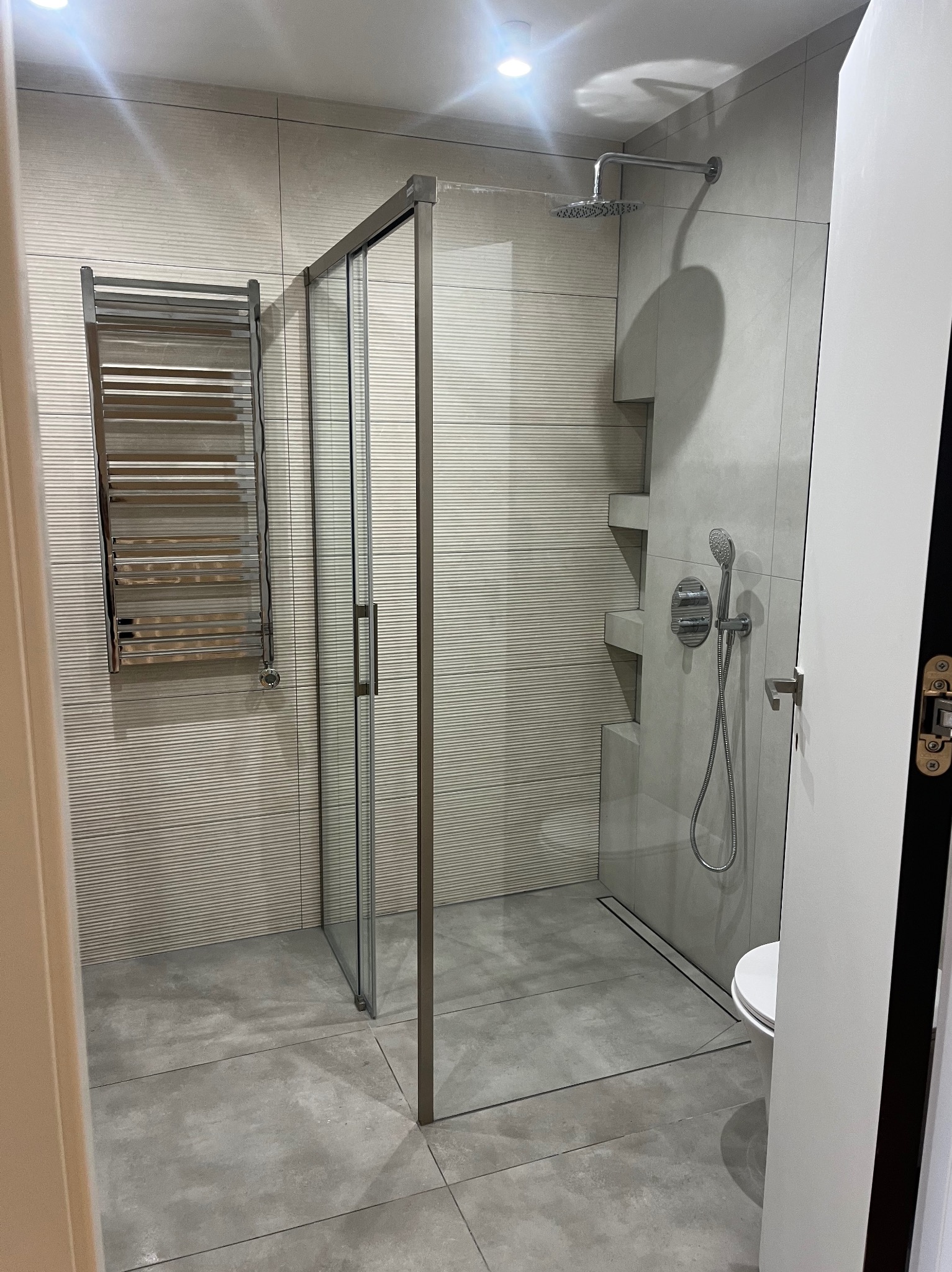 Nowoczesna łazienka z kabiną prysznicową typu walk-in, grzejnikiem drabinkowym i płytkami w odcieniach szarości z subtelną fakturą. Minimalistyczny design, widok zza otwartych drzwi.