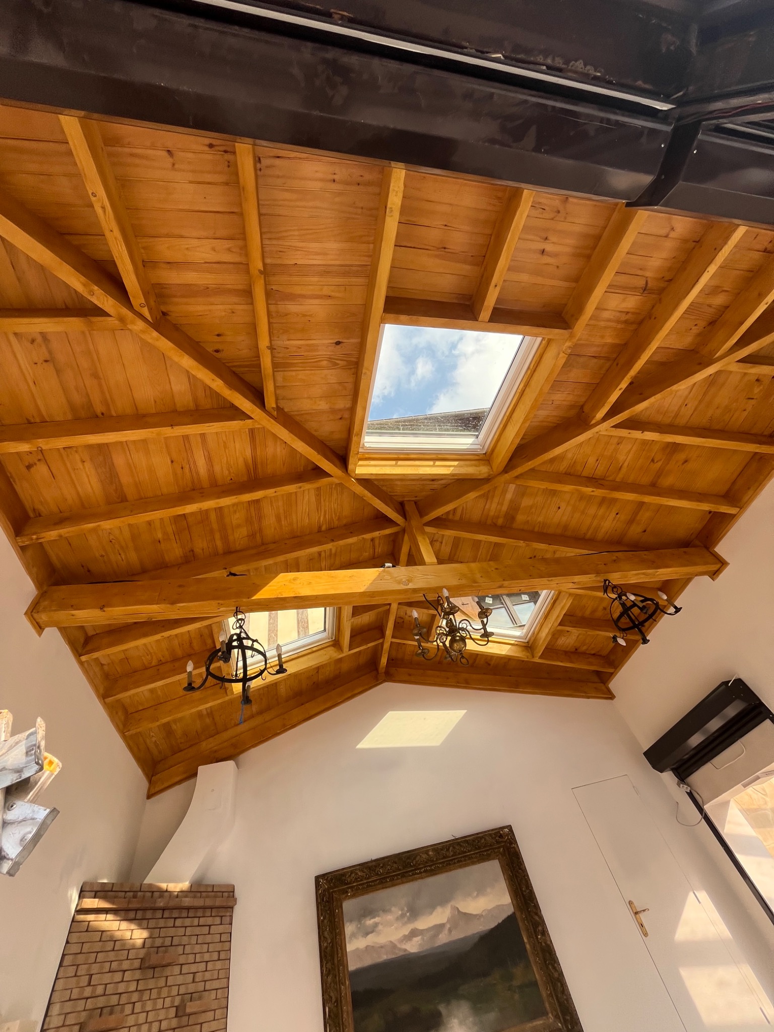 Drewniany sufit poddasza z oknami dachowymi i żyrandolami, widok z dołu. Jasne drewno, białe ściany, obraz w ozdobnej ramie. Nietypowy kąt ujęcia.