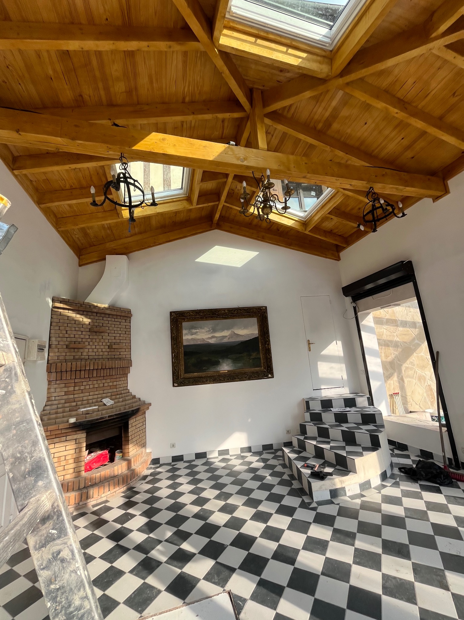 Jasne wnętrze z drewnianym stropem, oknami dachowymi i żyrandolami. Widoczny kominek z cegły, obraz na ścianie i schody w szachownicę. Częściowo widoczna drabina.