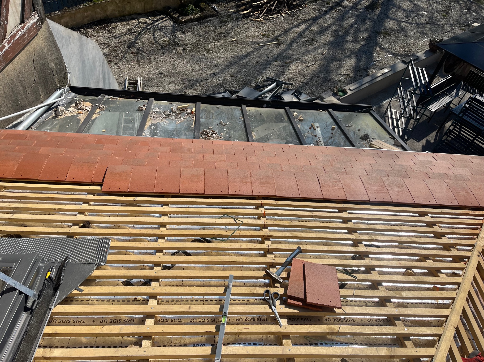 Montaż czerwonej dachówki na drewnianej konstrukcji dachu. Widoczne narzędzia i fragmenty starego dachu. Ujęcie z góry, w słoneczny dzień.