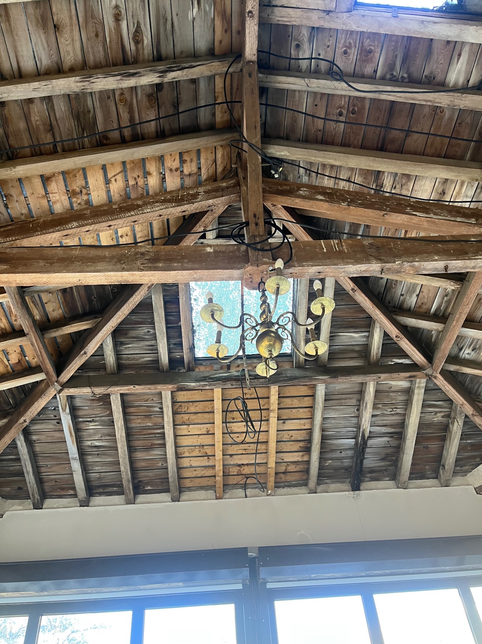 Drewniana konstrukcja dachu z widocznymi belkami, oknem dachowym i wiszącym żyrandolem. Widok z dołu, z fragmentem okna na dole kadru.