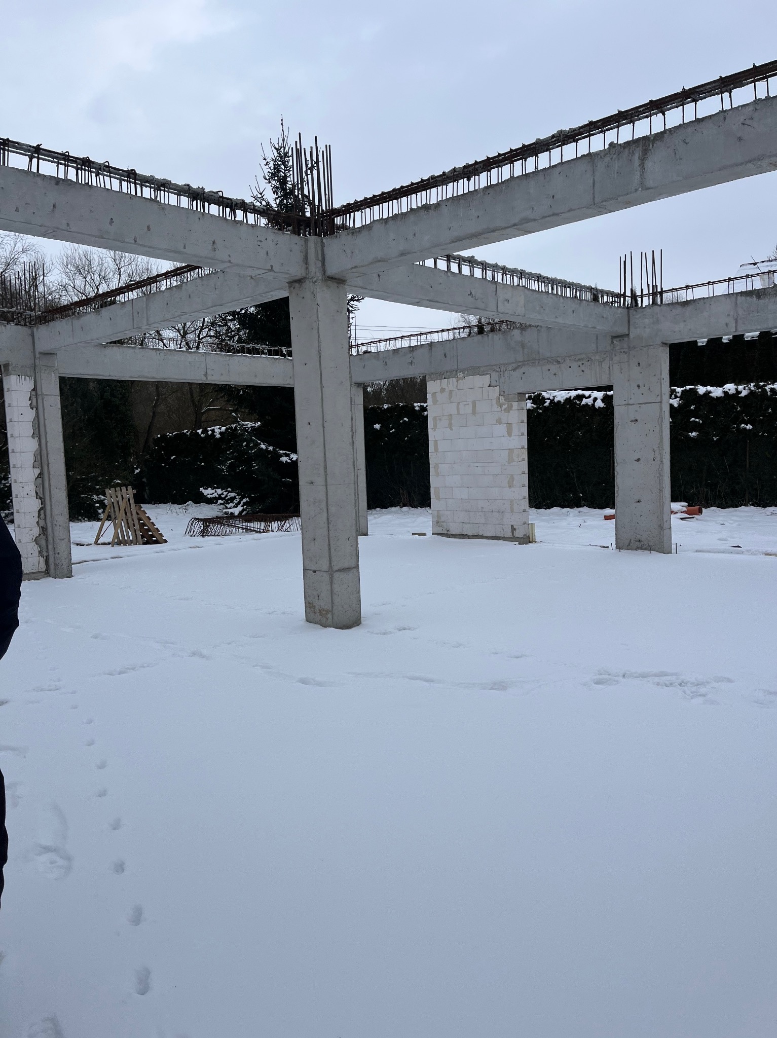 Surowa konstrukcja domu w zimowej scenerii: betonowe słupy i belki zbrojeniowe na tle śnieżnego krajobrazu. Częściowo wymurowane ściany z białych bloczków.