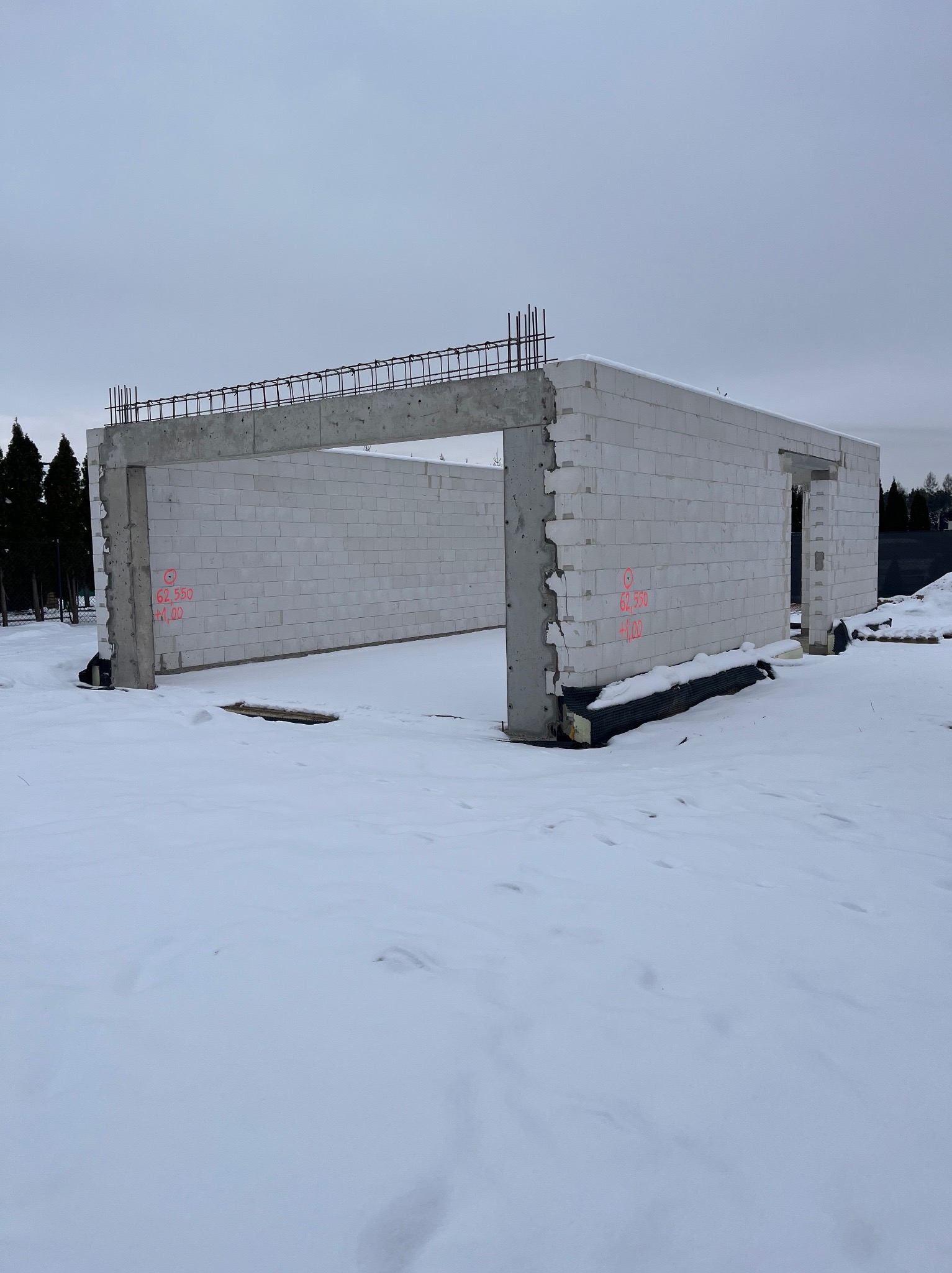 Surowy stan budynku z betonu komórkowego w zimowej scenerii. Widoczne zbrojenia na stropie i oznaczenia geodezyjne na ścianach. Fundament zabezpieczony izolacją.