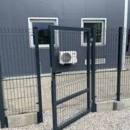 Otwarta furtka z szarej siatki panelowej na tle szarego budynku z dwoma oknami i klimatyzatorem. Furtka osadzona na betonowej podmurówce, podłoże wysypane białym żwirem.