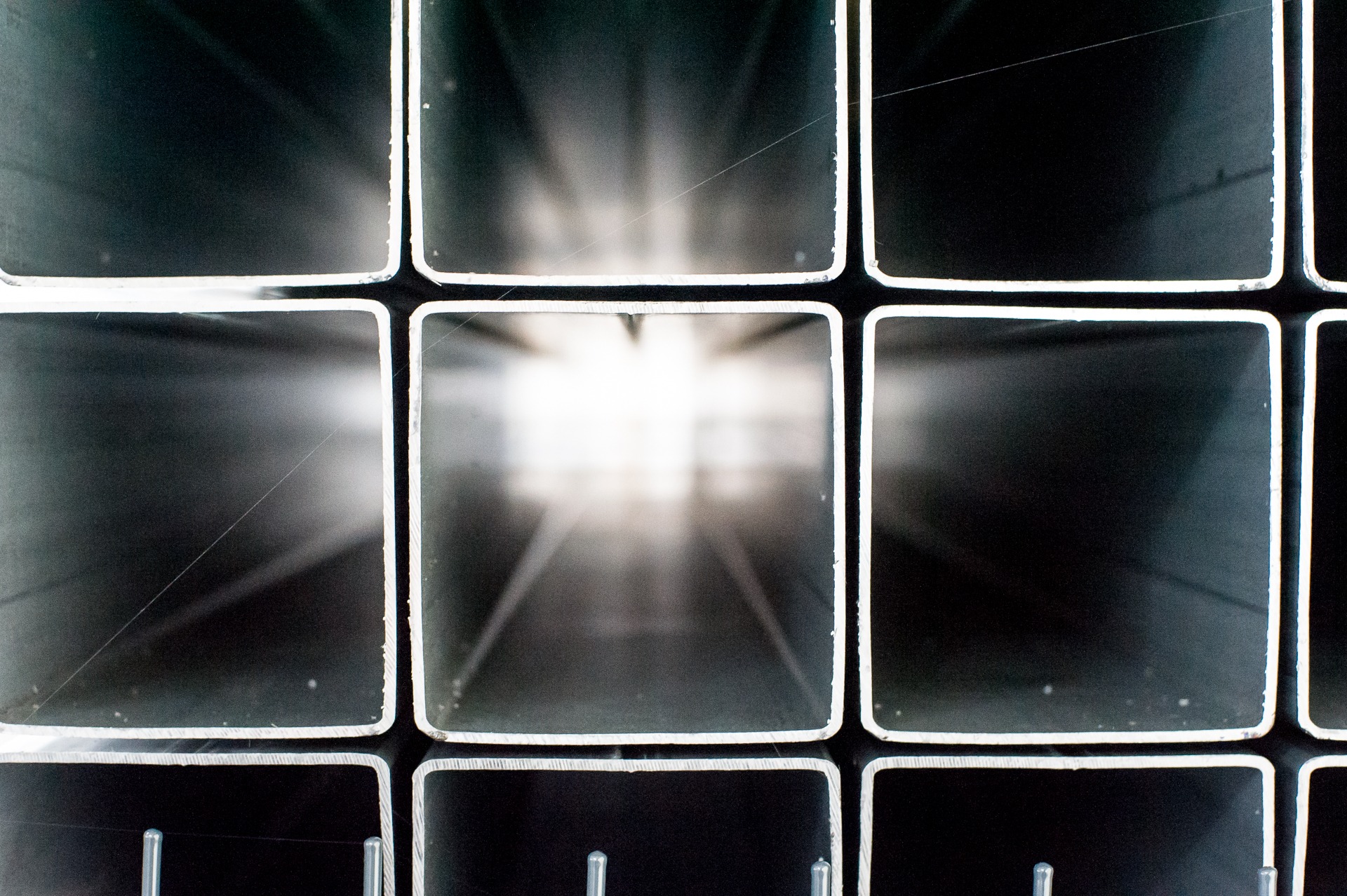 Metalowe profile kwadratowe ułożone w siatkę, z widocznym światłem przechodzącym przez środek, tworząc abstrakcyjny wzór. Widoczne drobne rysy na powierzchni metalu.