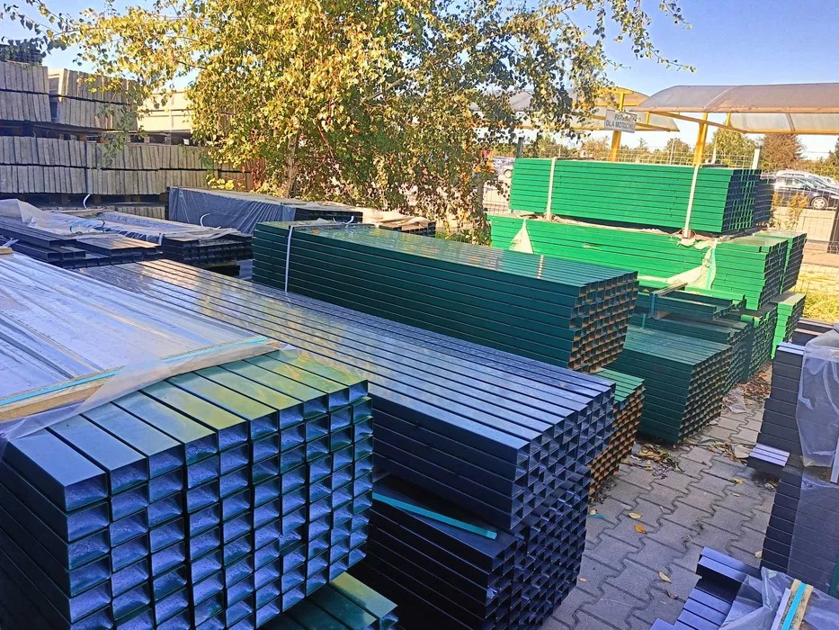 Stosy profili metalowych o przekroju kwadratowym, w odcieniach zieleni i szarości, składowane na zewnątrz, prawdopodobnie w magazynie lub składzie budowlanym.