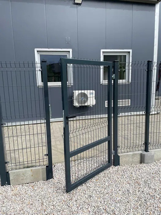 Otwarta furtka z szarej siatki panelowej na tle szarego budynku z dwoma oknami i klimatyzatorem. Furtka osadzona na betonowej podmurówce, podłoże wysypane białym żwirem.