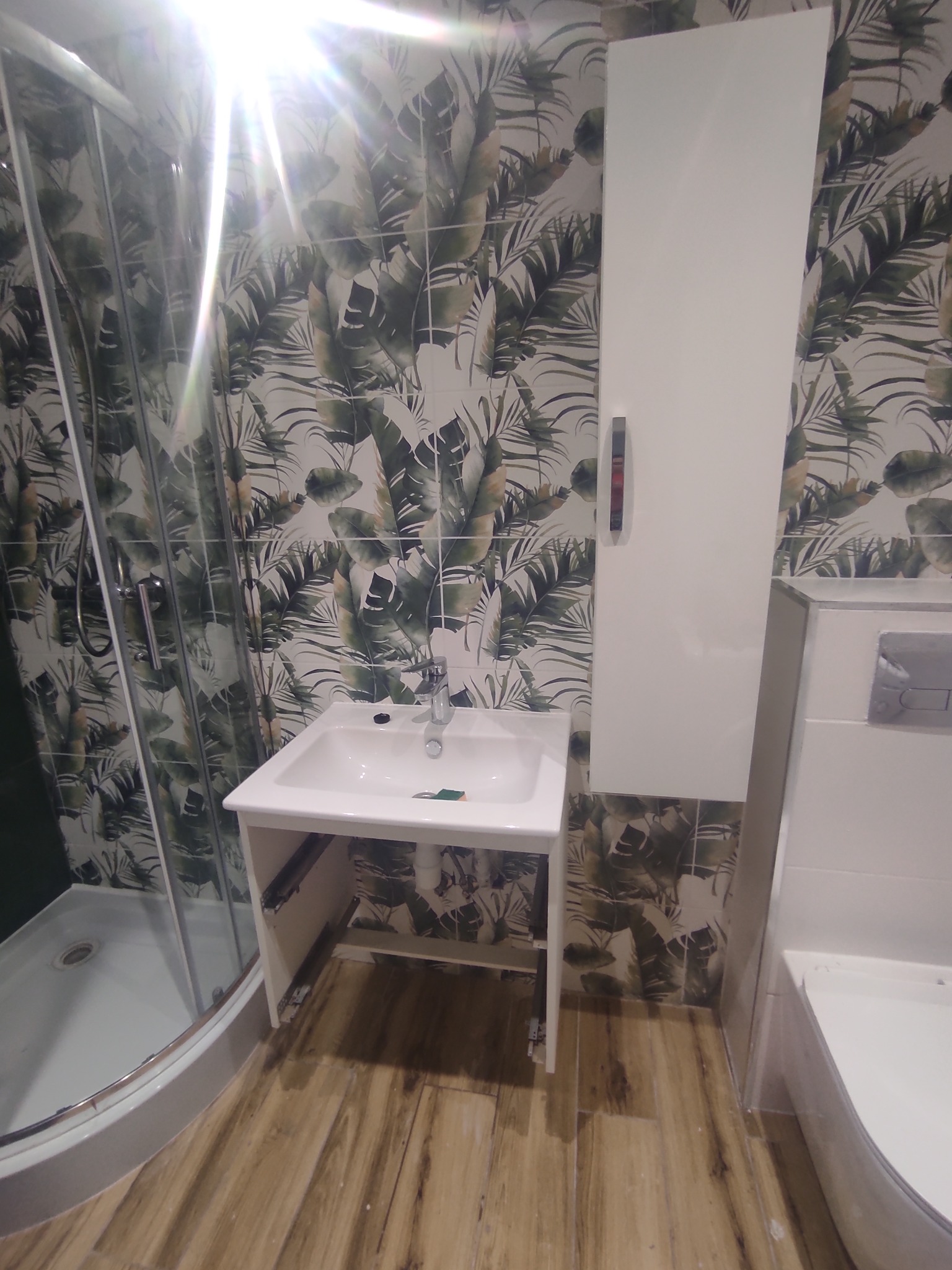 Ukończona łazienka z kabiną prysznicową, umywalką i szafką. Na ścianach płytki z motywem liści, na podłodze panele imitujące drewno. Widoczny fragment wanny.