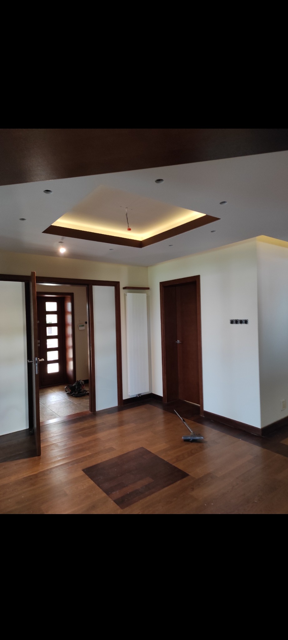 Nowoczesne wykończenie wnętrza: parkiet w odcieniach brązu, podwieszany sufit z oświetleniem LED i otwarte drzwi do kolejnych pomieszczeń. Widoczny mop na podłodze.