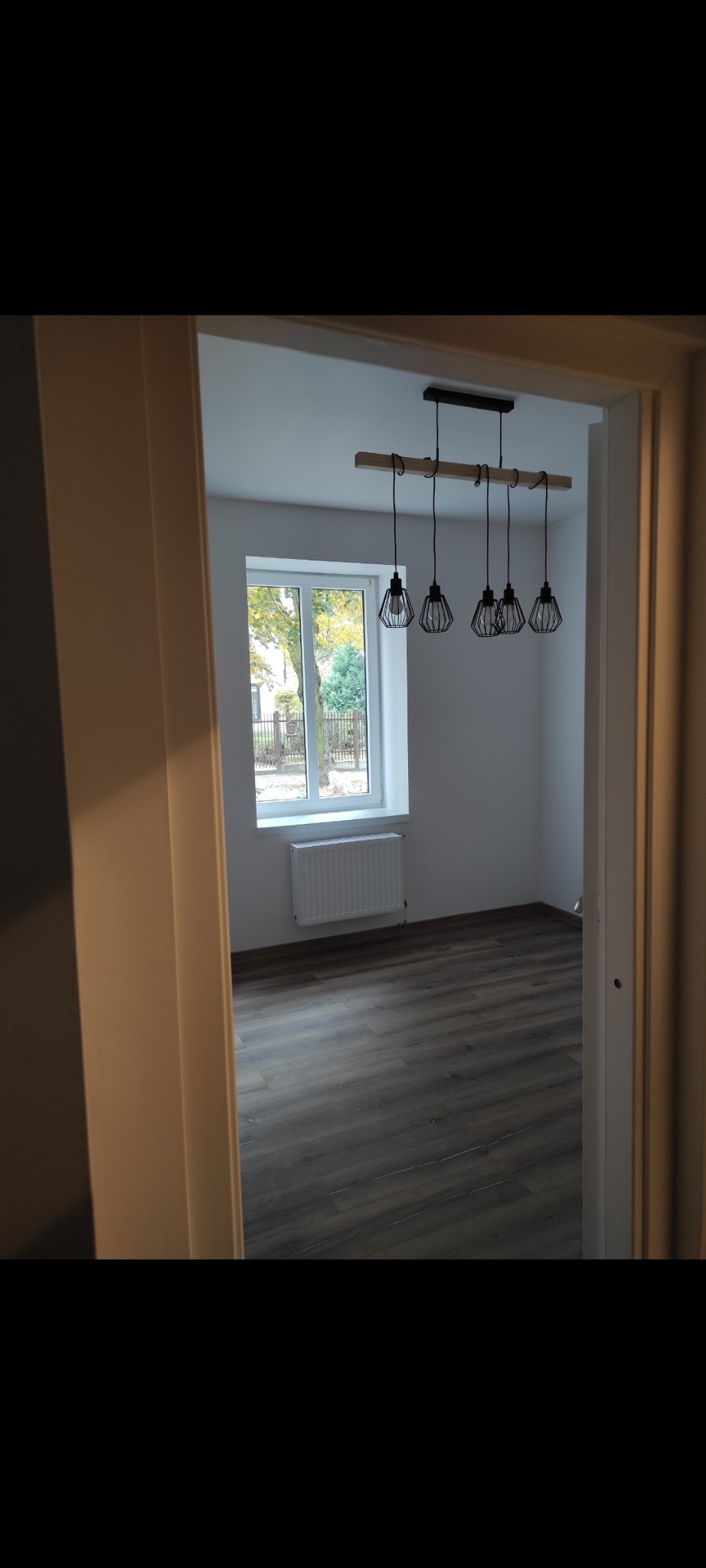 Wykończone wnętrze pokoju z drewnianą podłogą, białymi ścianami, oknem i nowoczesną lampą wiszącą z czarnymi kloszami, widoczne zza framugi drzwi.