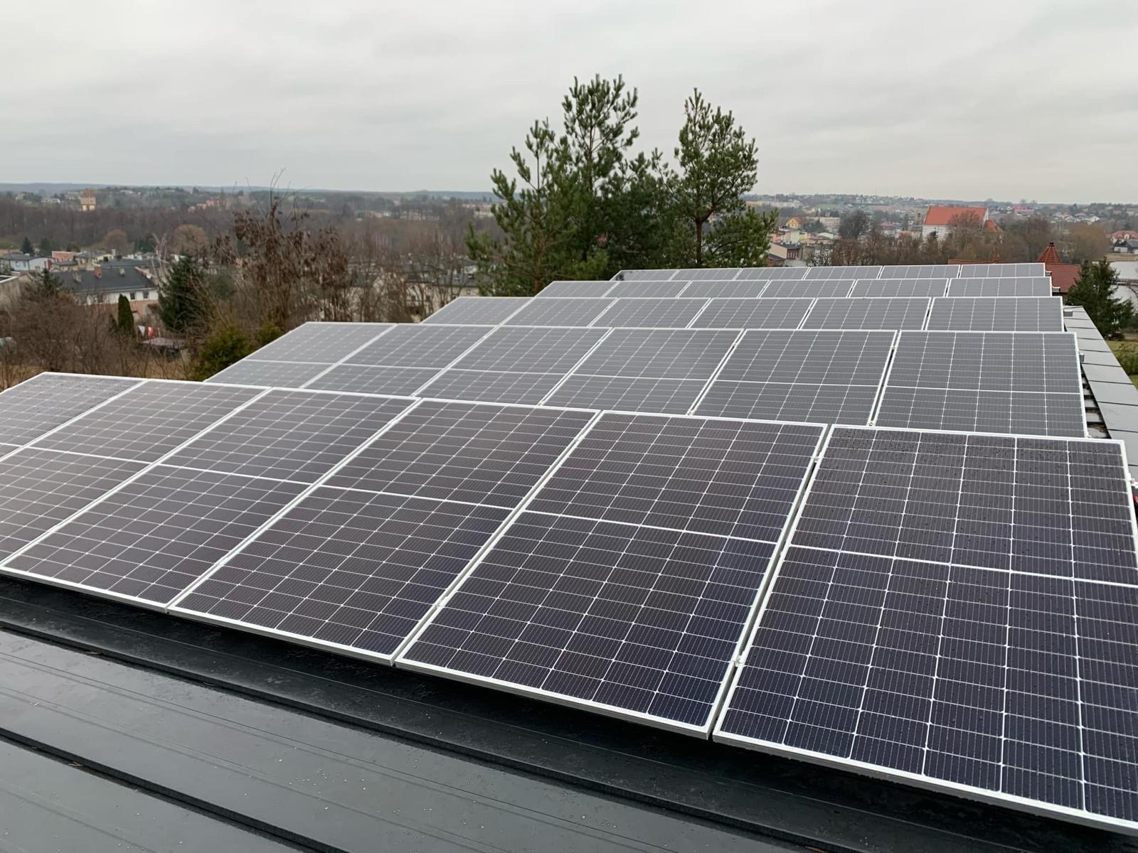 Rząd paneli fotowoltaicznych na dachu budynku, widok z góry, w tle zabudowa miejska i drzewa. Pochmurne niebo.