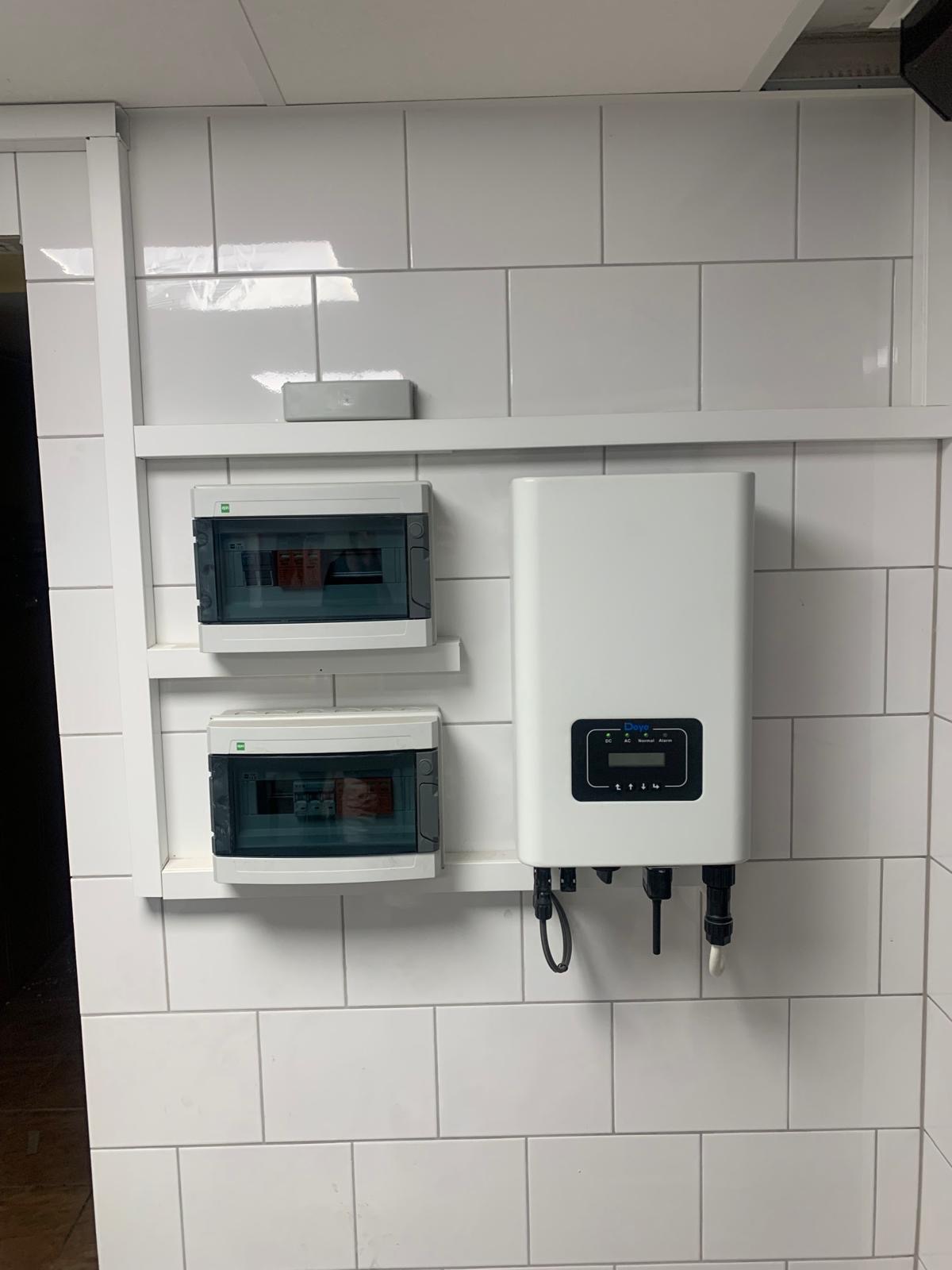 Biały inwerter fotowoltaiczny marki Daye zamontowany na ścianie z białych płytek, obok dwie skrzynki z bezpiecznikami na półce. Widoczne kable i złącza.