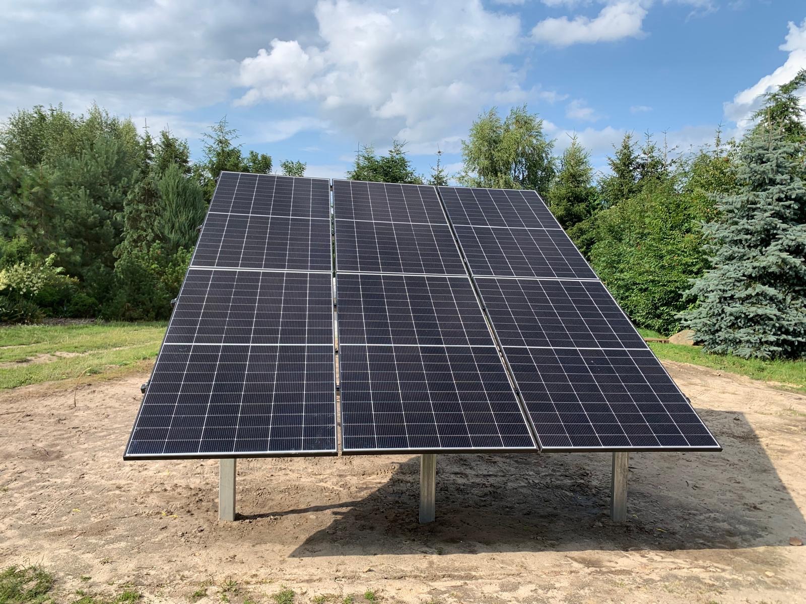 Panel fotowoltaiczny na metalowych podporach, zainstalowany na ziemi, z zielenią w tle i błękitnym niebem z chmurami. Widok z dołu, podkreślający solidność konstrukcji.