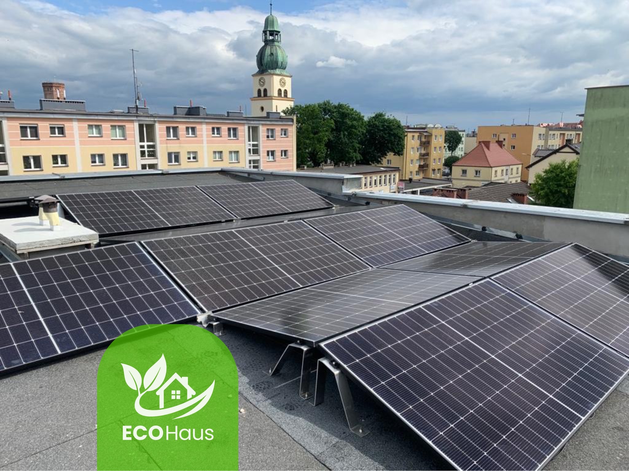 Instalacja paneli fotowoltaicznych na dachu budynku miejskiego, widok z góry, w tle wieża kościoła i zabudowa miejska, logo firmy w lewym dolnym rogu.