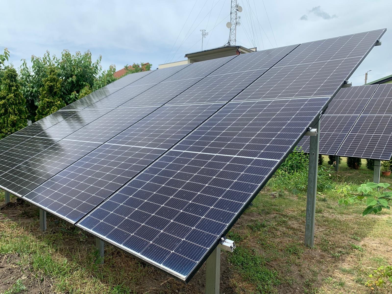 Instalacja fotowoltaiczna na gruncie, panele ustawione pod kątem, widoczne metalowe wsporniki, w tle budynek i maszt telekomunikacyjny.