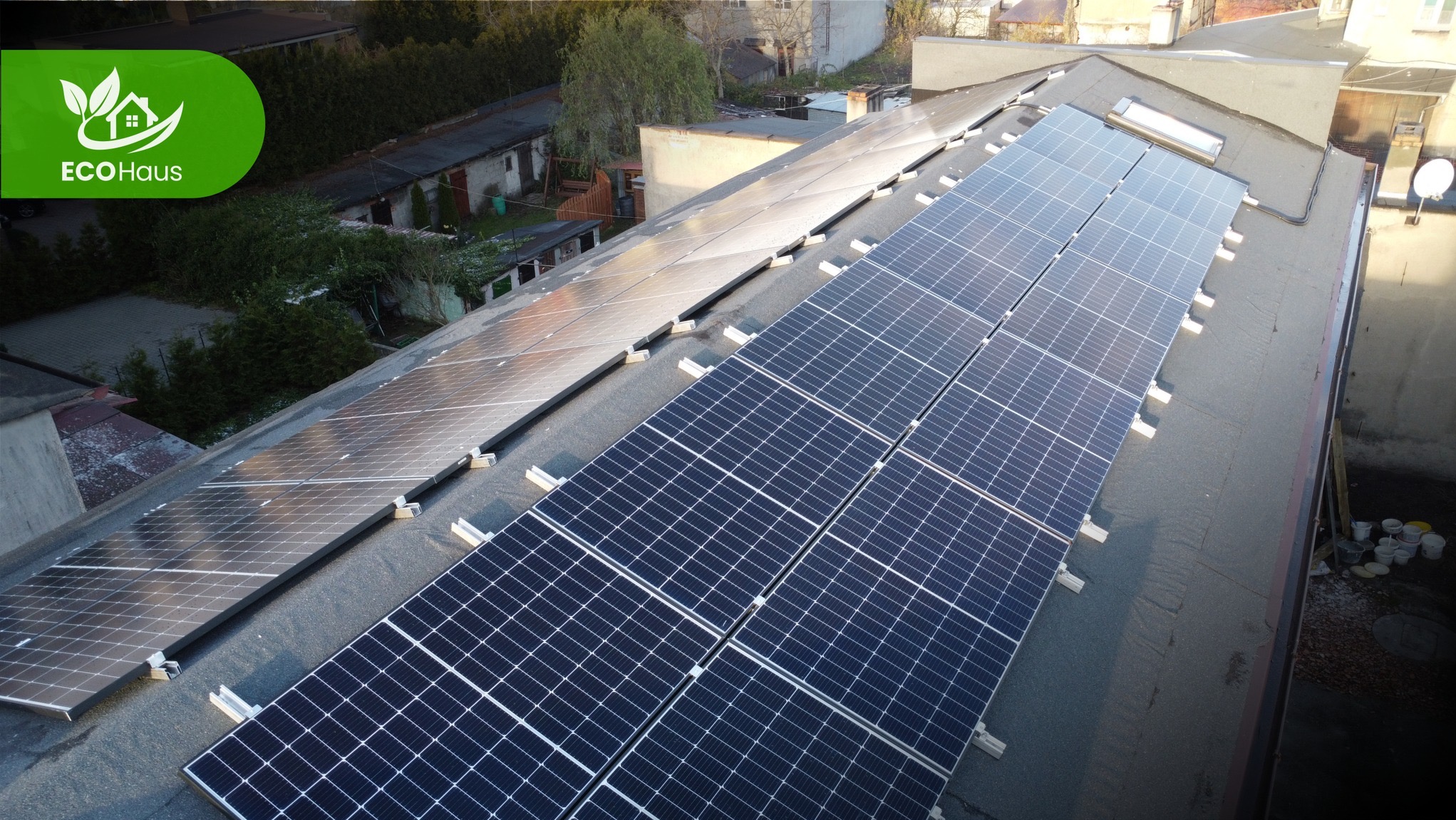 Panele słoneczne na dachu budynku z widocznymi elementami montażowymi. Ujęcie z góry, w tle zabudowa miejska i zieleń. Część paneli odbija światło.