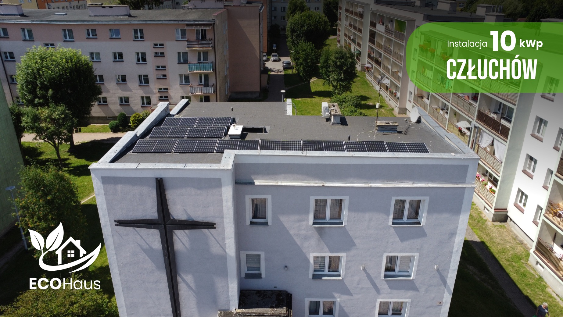 Instalacja fotowoltaiczna 10 kWp na dachu budynku z krzyżem, Człuchów. Logo EcoHaus w lewym dolnym rogu. Widok z góry na panele słoneczne.