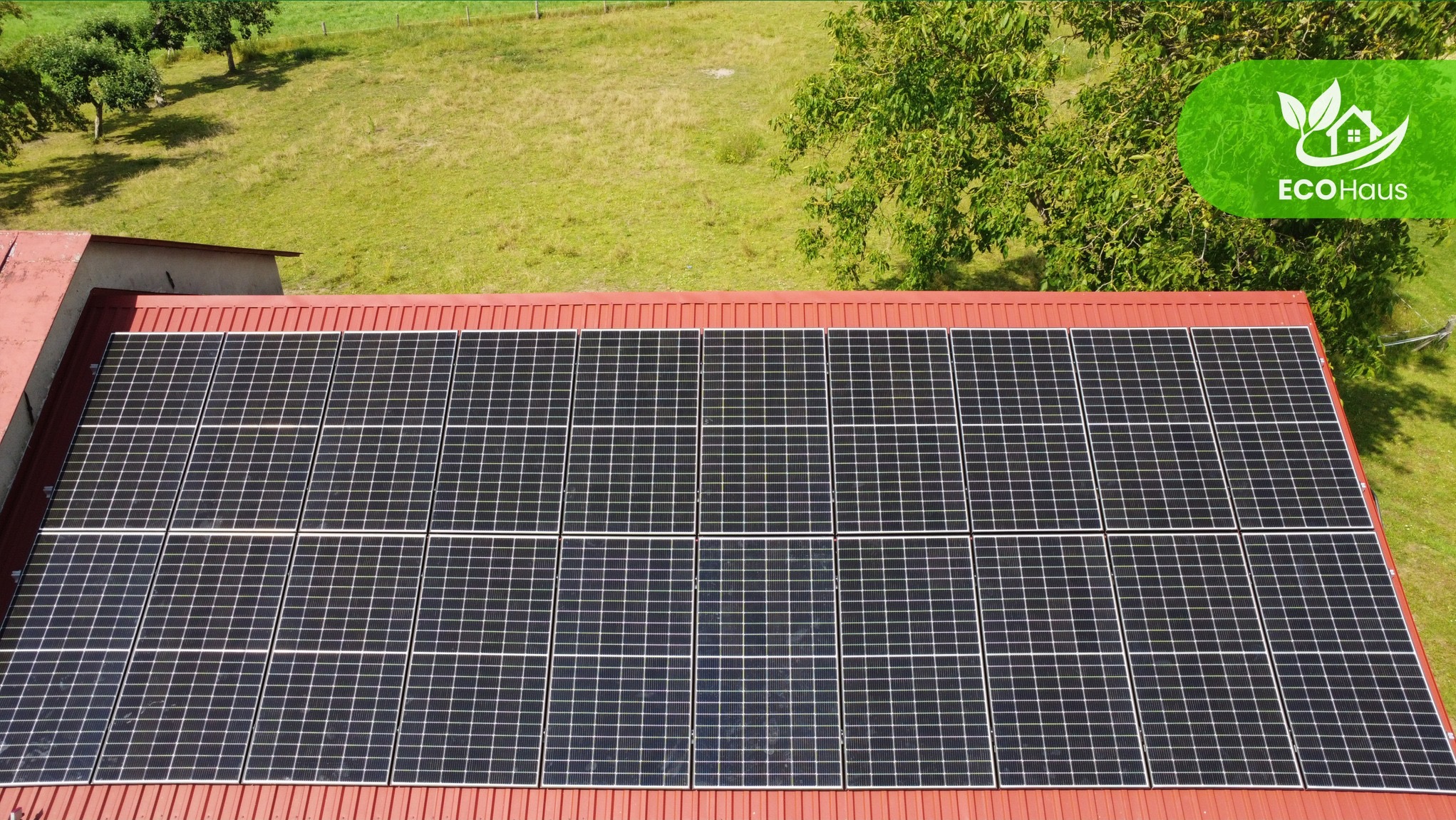 Widok z góry na panele fotowoltaiczne na czerwonym dachu budynku, otoczonego zielenią. Logo EcoHaus w prawym górnym rogu.