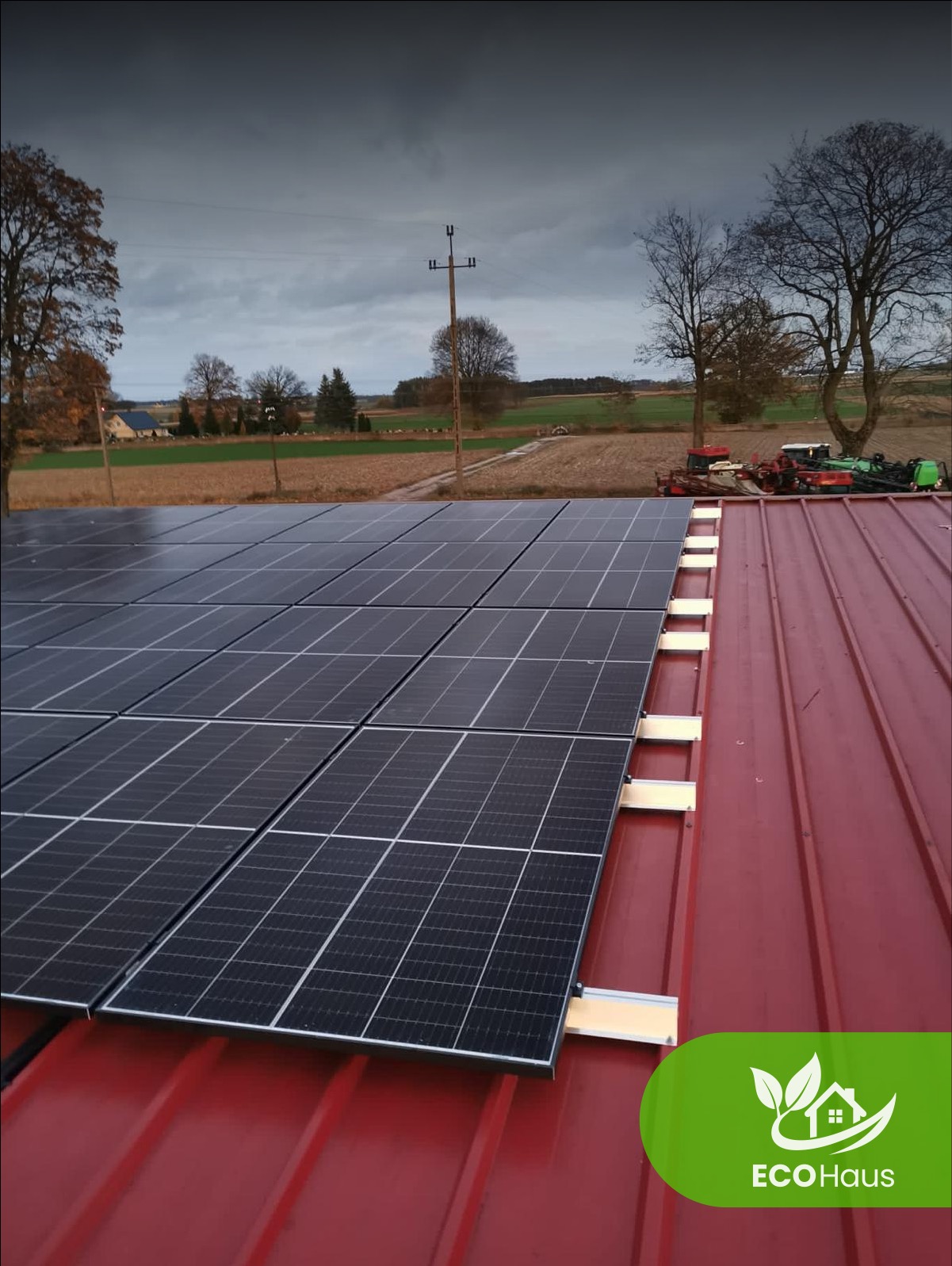 Panele słoneczne na czerwonym dachu, widok z góry, w tle pole i drzewa. Montaż paneli fotowoltaicznych w wiejskim otoczeniu.