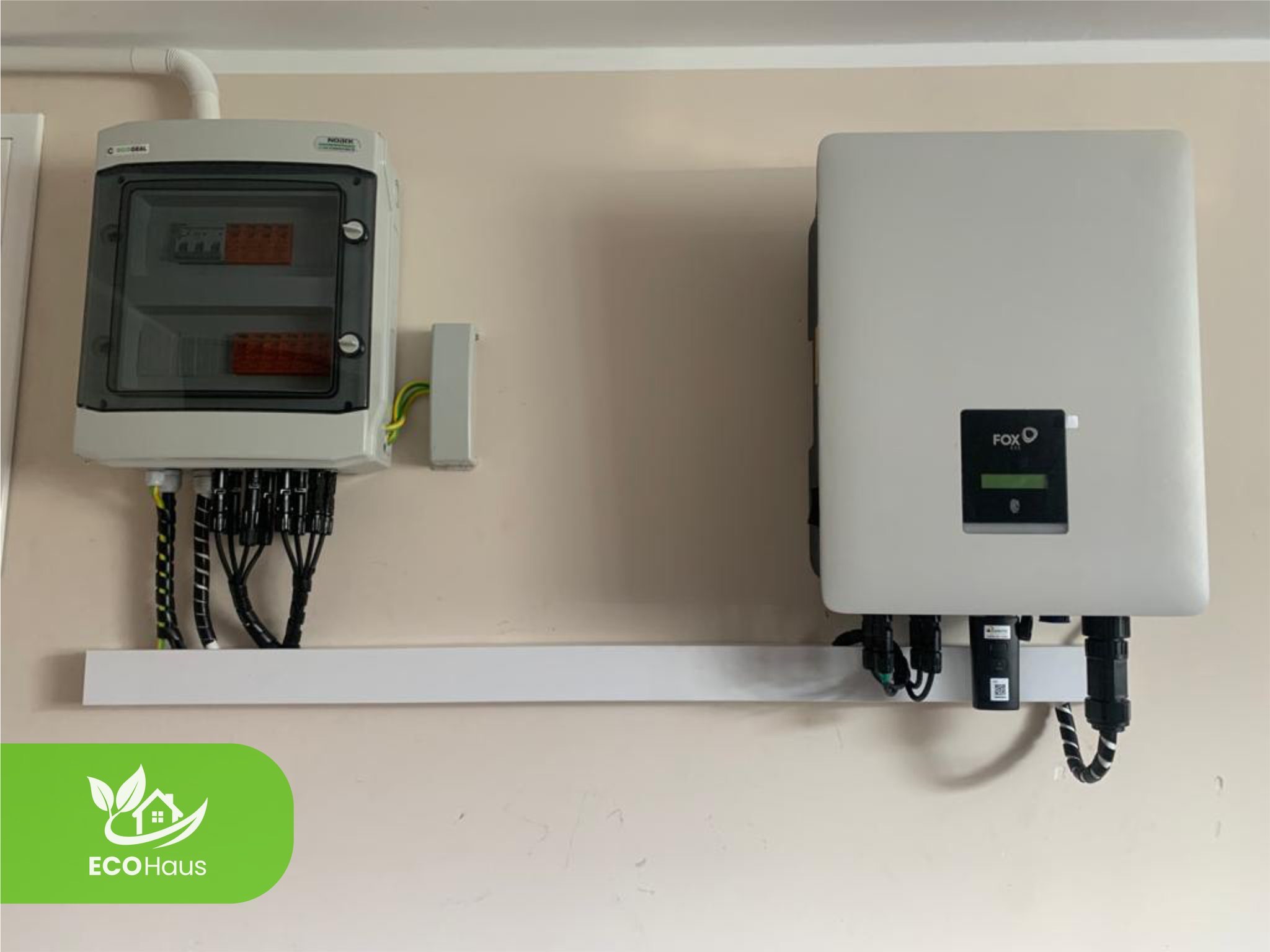 Zamontowana skrzynka rozdzielcza i falownik FOX na ścianie, połączone przewodami w peszlach. Biała listwa maskująca pod urządzeniami. Widoczne logo EcoHaus.