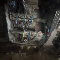 OSKAR WOŹNY - Rozdzielnica elektryczna z podłączonymi przewodami w różnych kolorach, widoczna przez przezroczystą obudowę. Element instalacji elektrycznej wewnątrz skrzynki.
