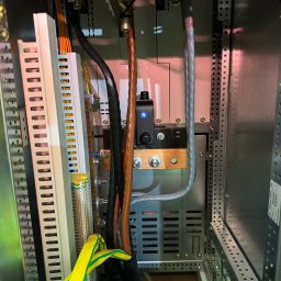 OSKAR WOŹNY - Wnętrze szafy elektrycznej z widocznymi przewodami, listwami zaciskowymi i elementami montażowymi. Kabel ochronny żółto-zielony. Widoczne złącza i zabezpieczenia.