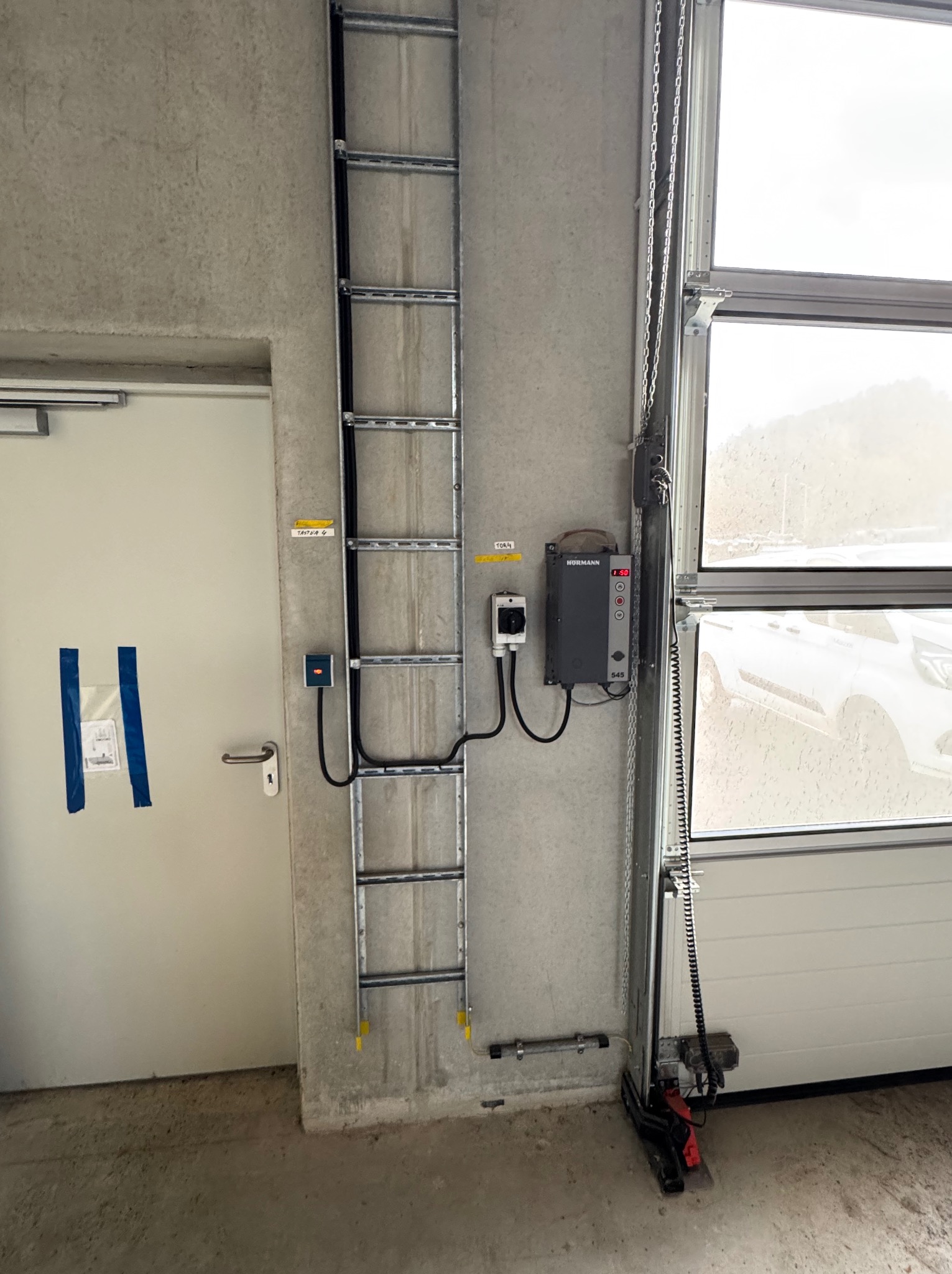 Szara skrzynka sterująca Hormann 545 z przyciskami i wyświetlaczem, zamontowana na betonowej ścianie obok metalowej drabinki kablowej i bramy garażowej.