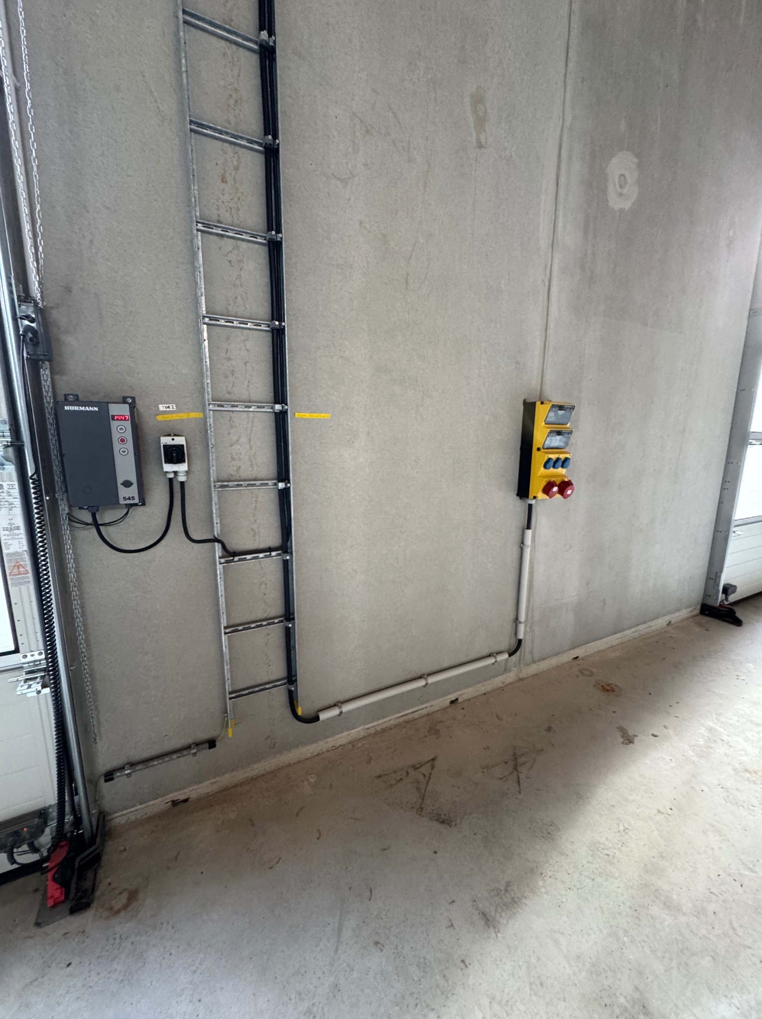 Szara ściana z zamontowaną drabinką, panelem sterowania i skrzynką elektryczną Hormann, prawdopodobnie w garażu lub hali przemysłowej. Widoczne elementy instalacji.