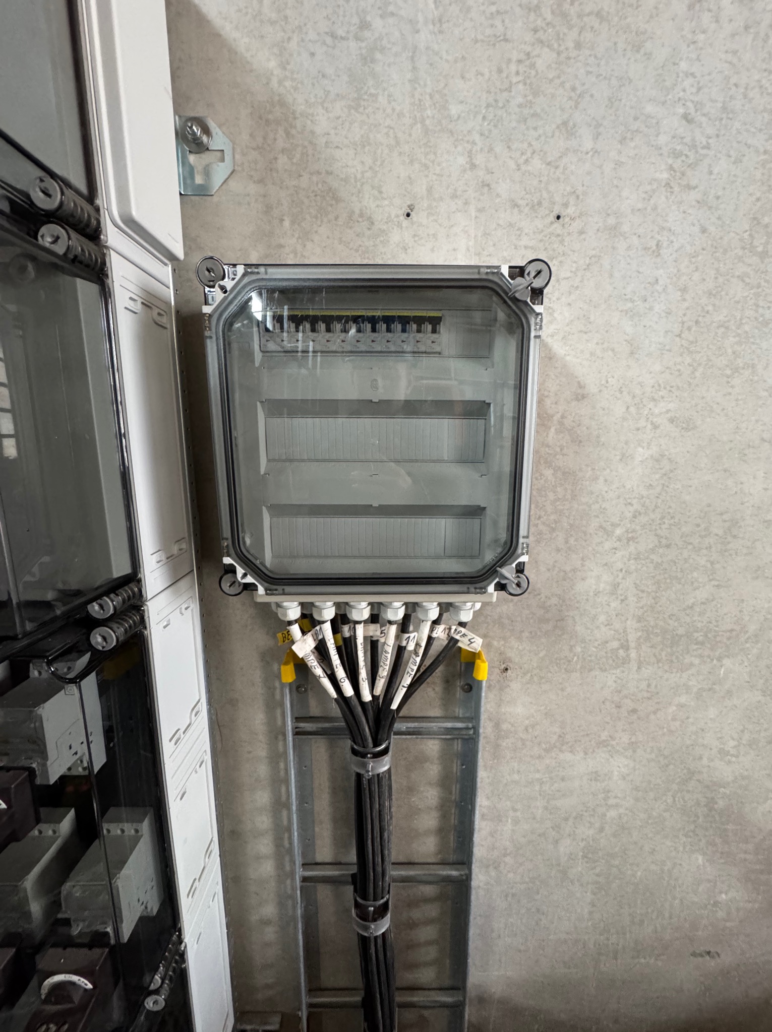 Szara skrzynka elektryczna z przezroczystym wiekiem, przymocowana do ściany, z podłączonymi kablami oznaczonymi etykietami, widok z bliska.