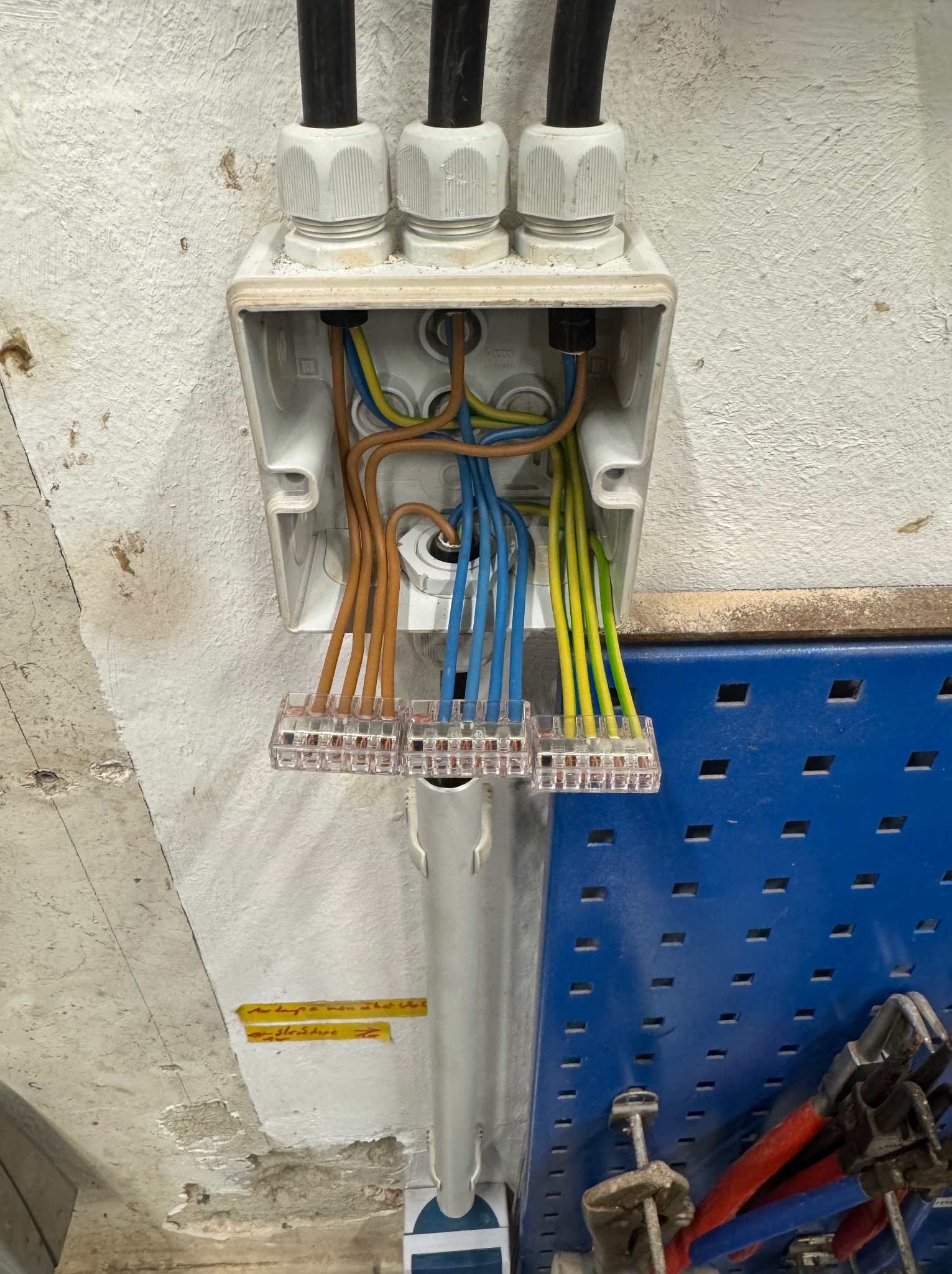 Otwarta puszka elektryczna z podłączonymi przewodami o różnych kolorach, zamocowana na nierównej ścianie. Widoczne złącza i narzędzia na niebieskiej tablicy.