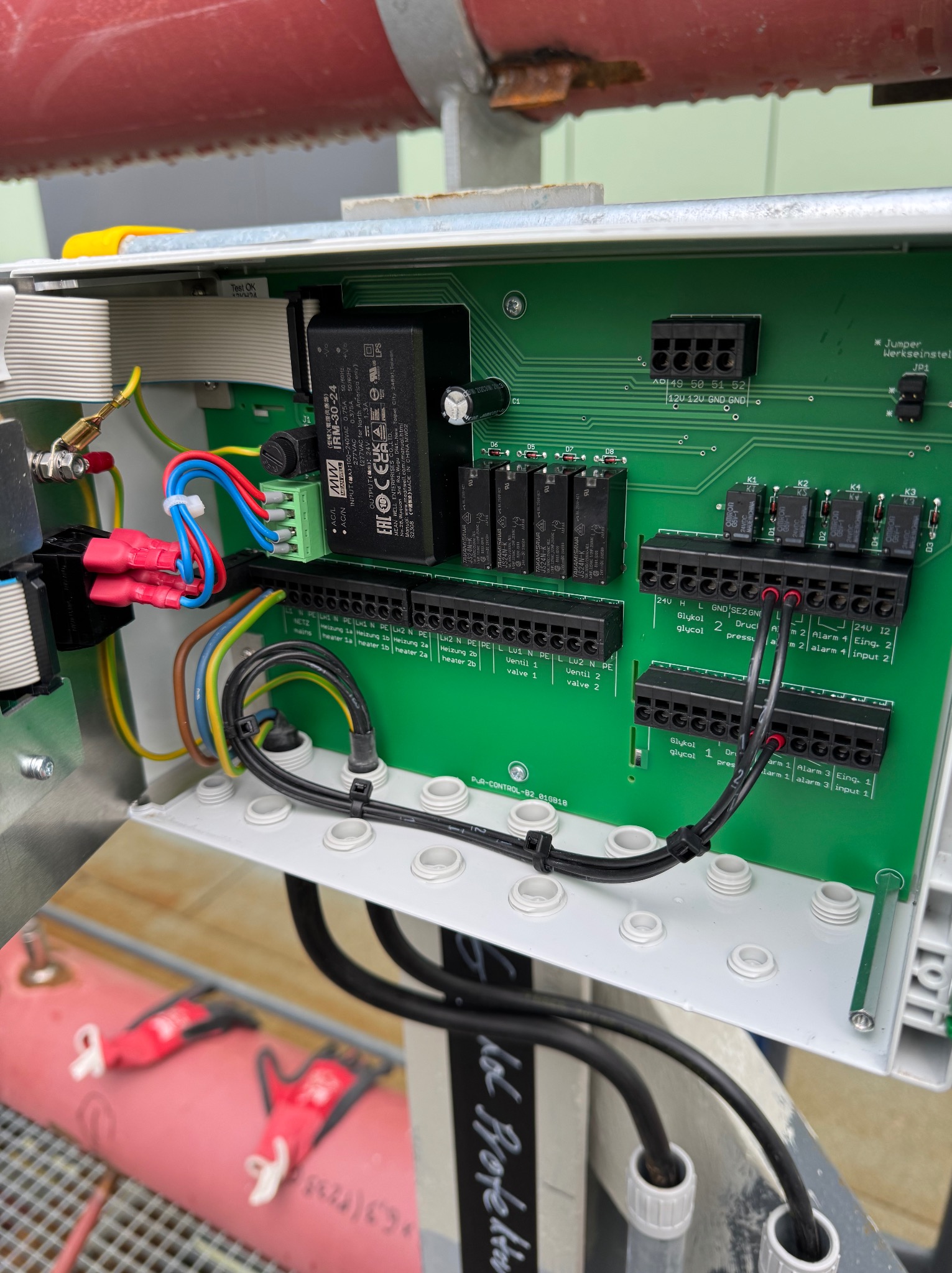 Otwarta skrzynka sterownicza z zieloną płytą drukowaną, widoczne liczne złącza, kable i przekaźniki. Elementy instalacji elektrycznej wewnątrz obudowy.