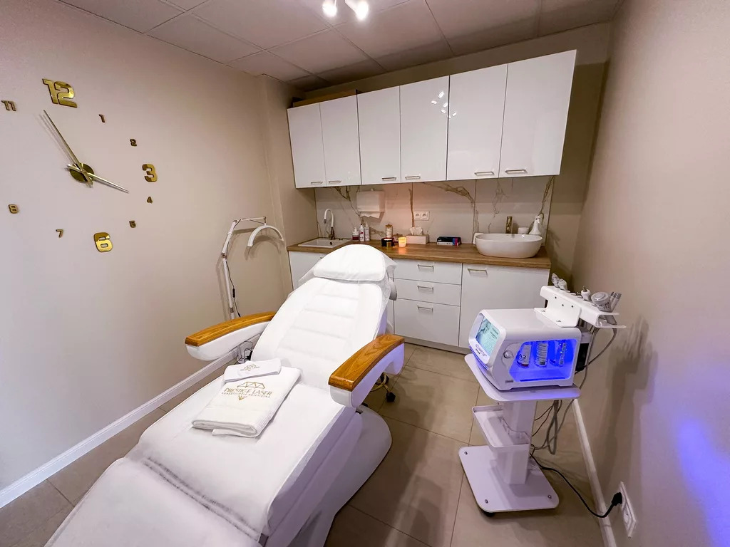 Jasne wnętrze salonu kosmetycznego z białym fotelem zabiegowym, nowoczesnym sprzętem i złotym zegarem ściennym. Widok na umywalkę i szafki.