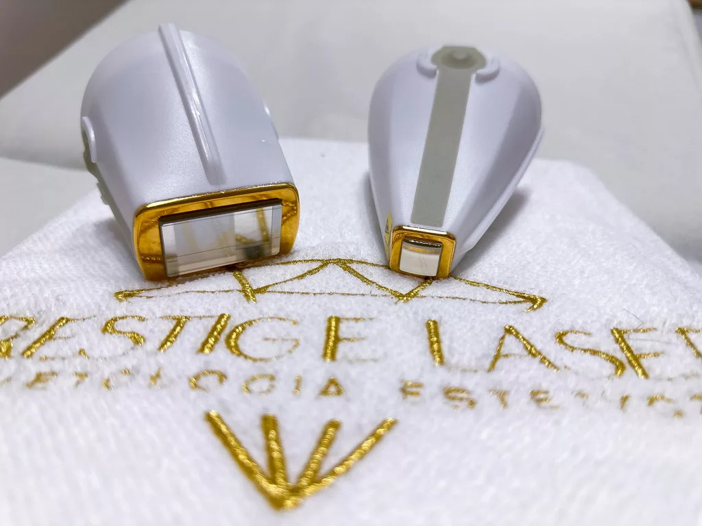 Dwie głowice lasera do depilacji, biało-złote, leżą na białym ręczniku z wyhaftowanym złotym napisem 'Prestige Laser'.
